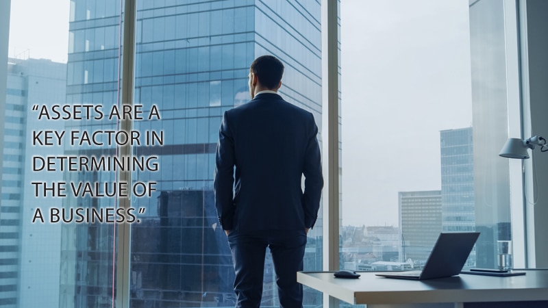 Back View of the Thoughtful Businessman wearing a Suit Standing in His Office, Hands in Pockets and Contemplating Next Big Business Deal, Looking out of the Window. Big City Business District Panoramic Window View.